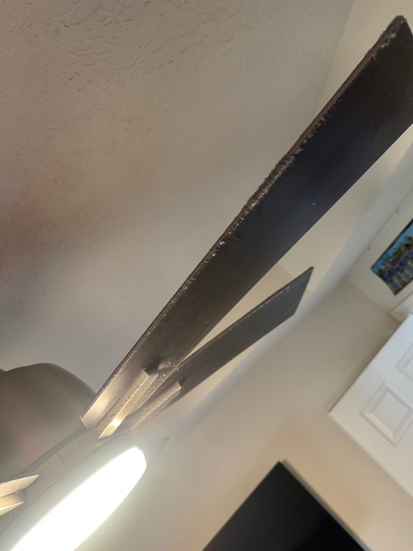 Looking up at a dark ceiling fan blade before cleaning, with a thick line of dust caked along its leading edge.