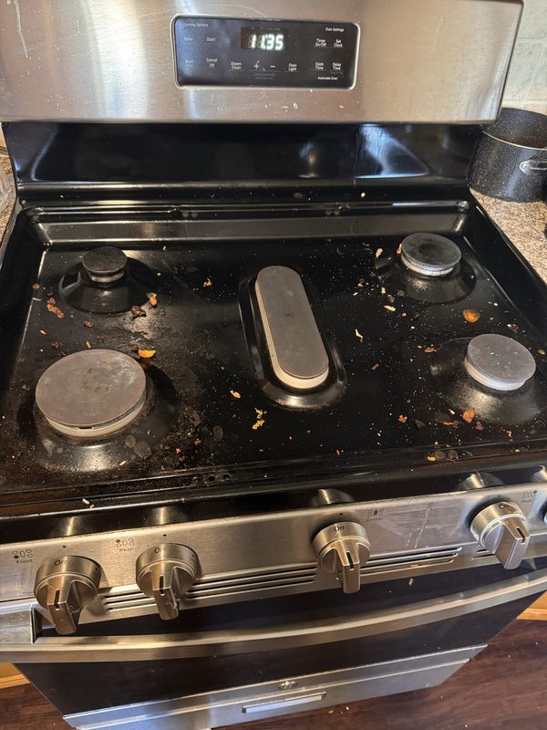 A gas cooktop before cleaning, with crumbs, dried food and grease spatter scattered across the black surface and around the burner caps.