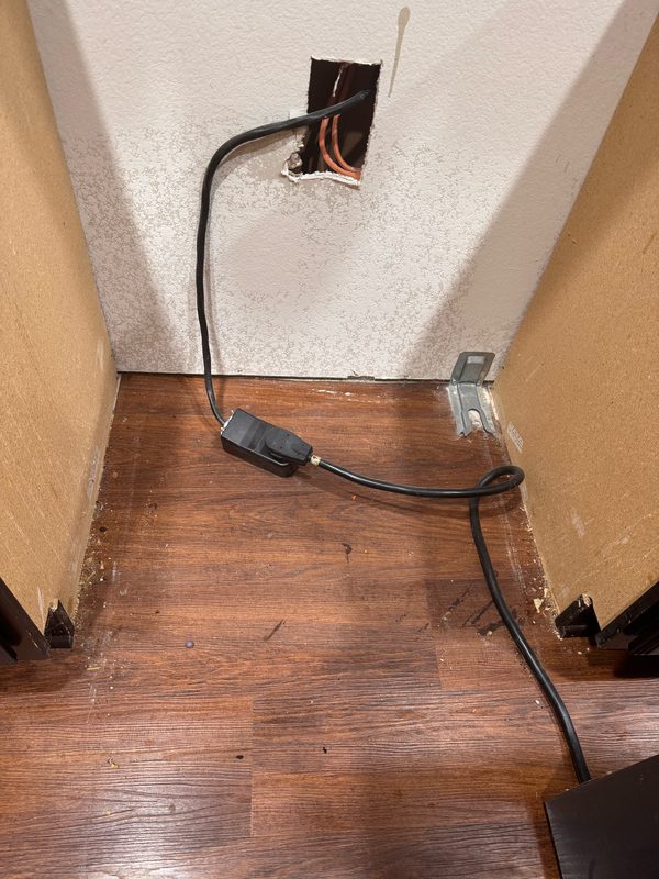 The floor in the gap behind a pulled-out appliance before cleaning, with crumbs and grit collected along the seam where the wood floor meets the wall.