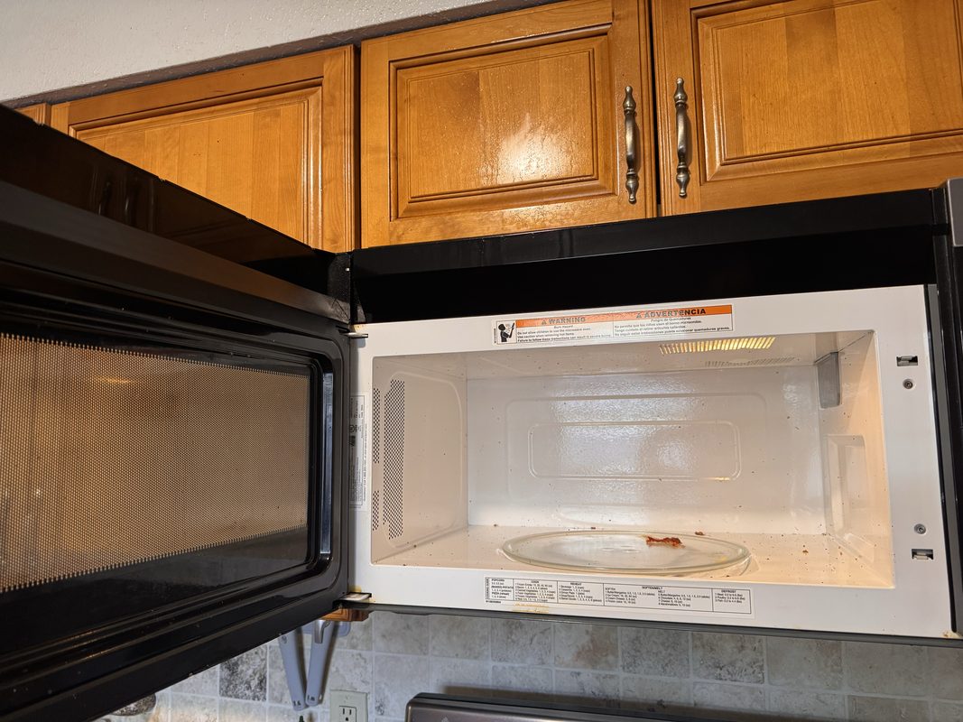 The open interior of an over-the-range microwave before cleaning, with food spatter dried onto the glass turntable and specks across the cavity floor and walls.