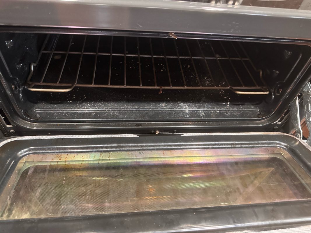 An open oven before cleaning, with baked-on spatter across the inside of the door glass and grease film down the cavity walls.