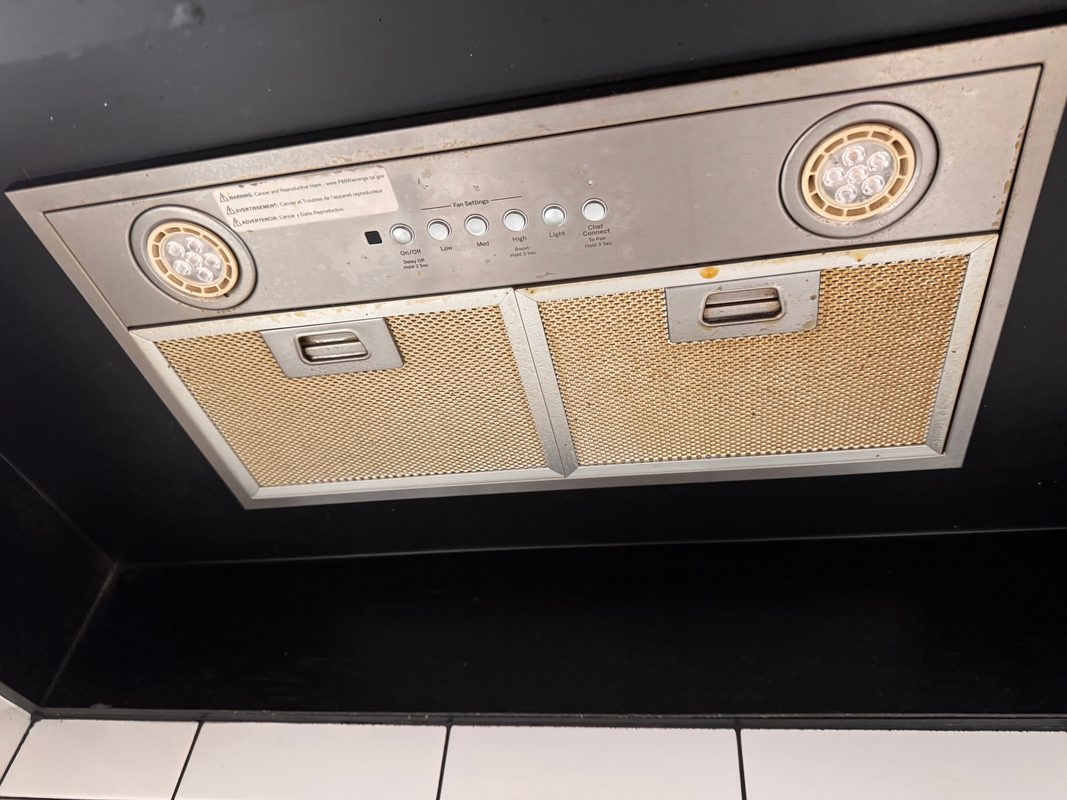 The underside of a stainless range hood before cleaning, seen from below against a black cabinet, with both mesh filters stained amber with grease.
