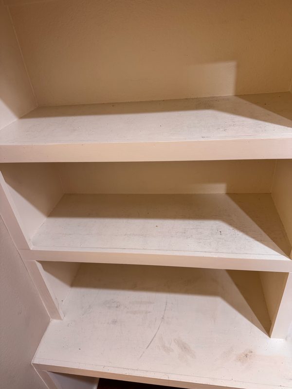 Three stacked shelves in a white built-in cupboard before cleaning, with grey scuff marks and smudges across each shelf surface.