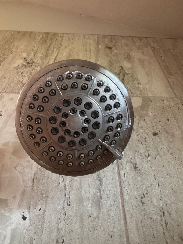 A round shower head seen from below before cleaning, with white buildup crusted around its rim and over many of its nozzles, against grey stone-look tile.