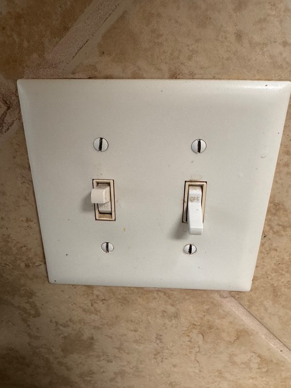 A white double toggle switch plate on a beige tile wall before cleaning, yellowed with grime and darkened around both toggles.