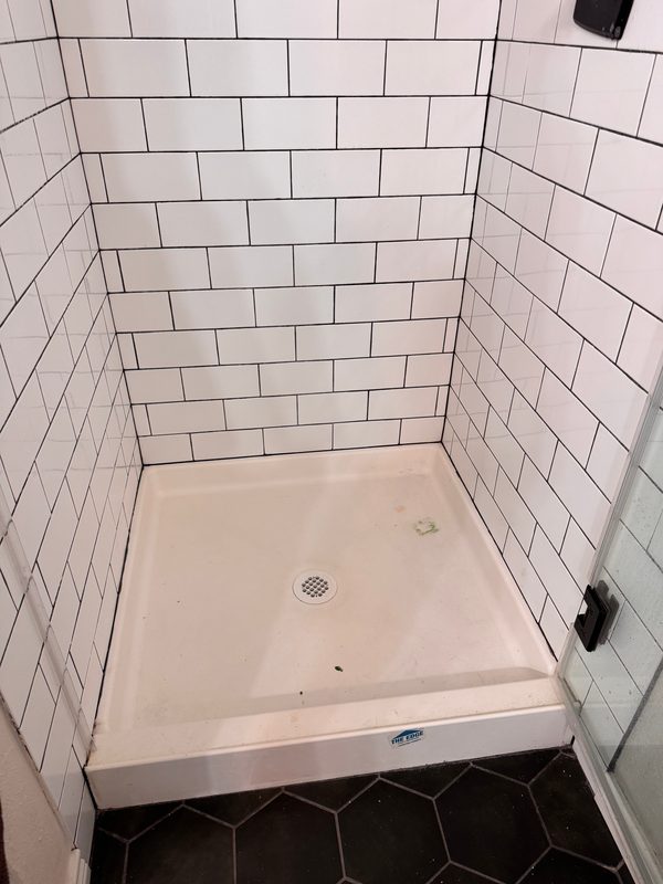 A walk-in shower with white subway tile and a white shower pan before cleaning, seen from the open door, with hair and specks of debris across the pan.
